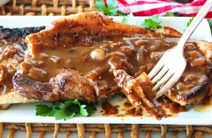 Pork steaks on a platter, smothered with gravy. A bite has been cut out and the fork is picking it up and holding it toward the camera.