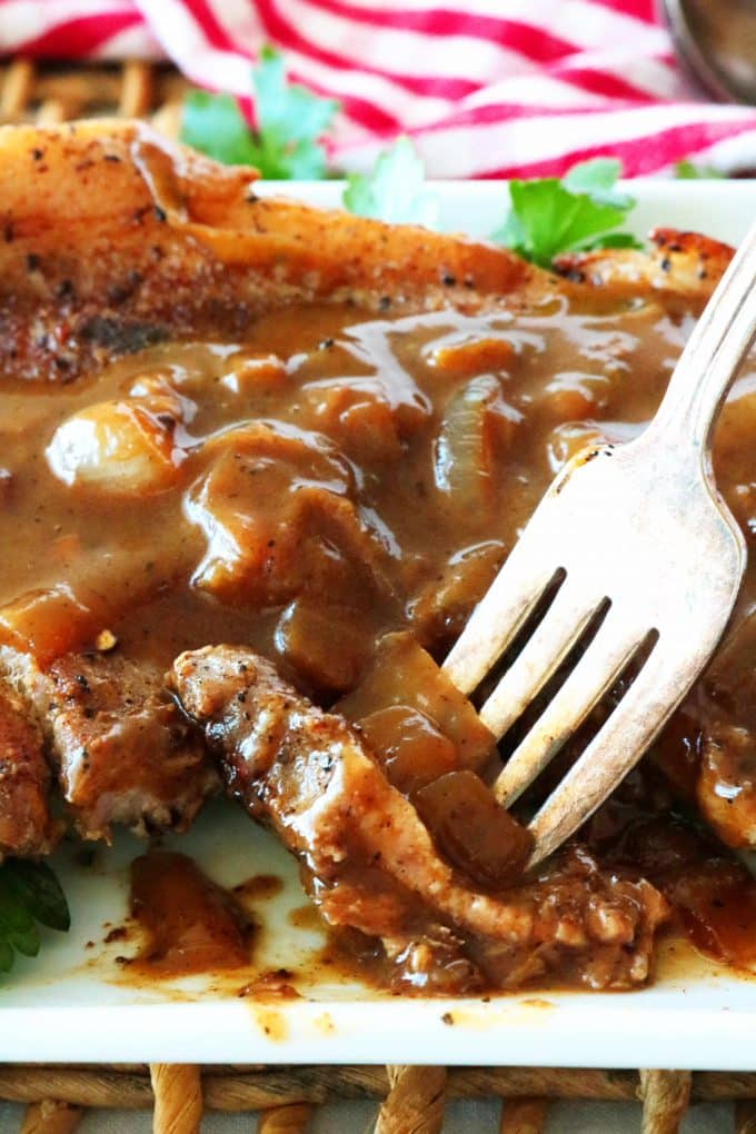 Pork steak smothered with brown onion gravy on a plate with a fork holding a bite cut out of it and slightly lifting it up.