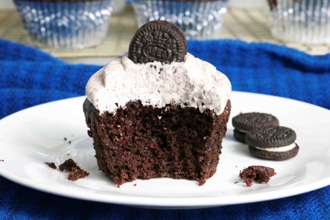 A bit missing from an Oreo Cupcakes that is served on a white plate with a couple of mini oreos lying next to the cupcake. 
