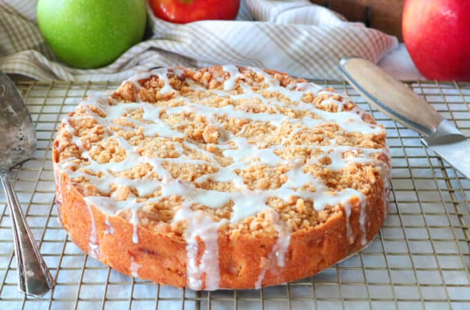 A round Apple Crumble Cake sitting on top of a wire cooling rack with whole red and green apples in the background. 