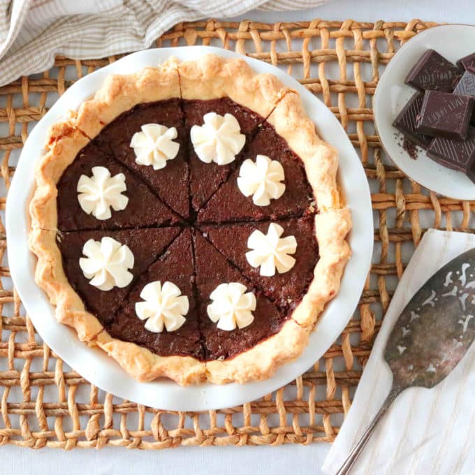 An overhead shot of Chocolate Chess Pie sitting on top of a woven wood placemat. Sitting next to the pie is a pie server and a plate of chocolate squares. 