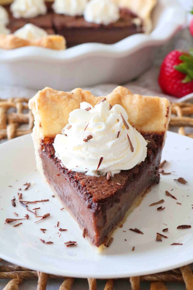 One slice of Chocolate Chess Pie on white plate. In the background is the rest of the pie in a scalloped pie dish. 