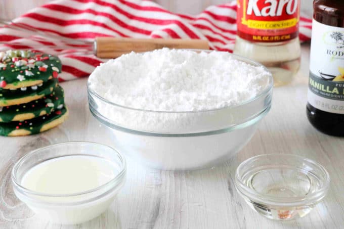 Ingredients for sugar cookie icing -- bowl of milk, powdered sugar, and corn syrup with bottles of vanilla and corn syrup in the background.
