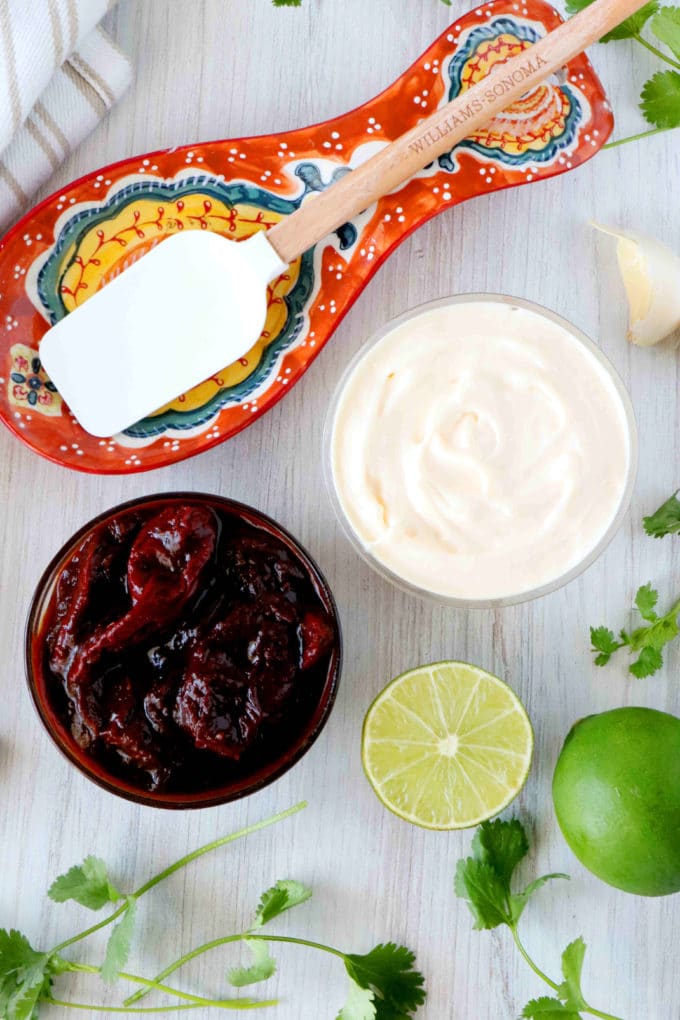 An overhead shot of a bowl of chipotle peppers in adobo sauce, cut up limes, and mayonnaise.