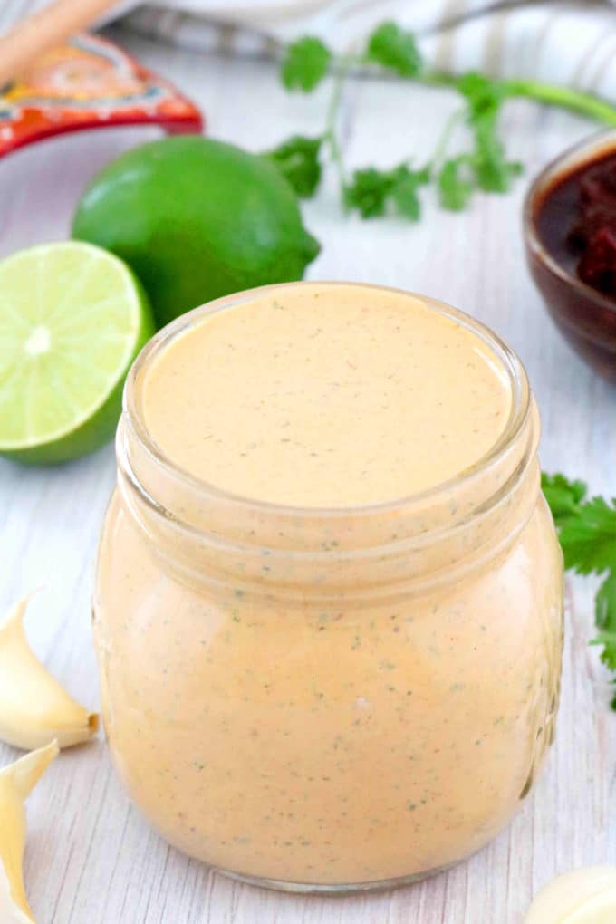 Chipotle sauce in a jar with limes cut up behind it, sprigs of cilantro, and a small bowl of adobo sauce.