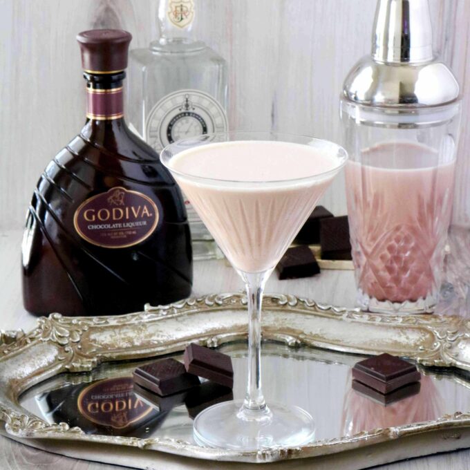 A martini on a glass tray with bottles of liquor and and shaker behind it.