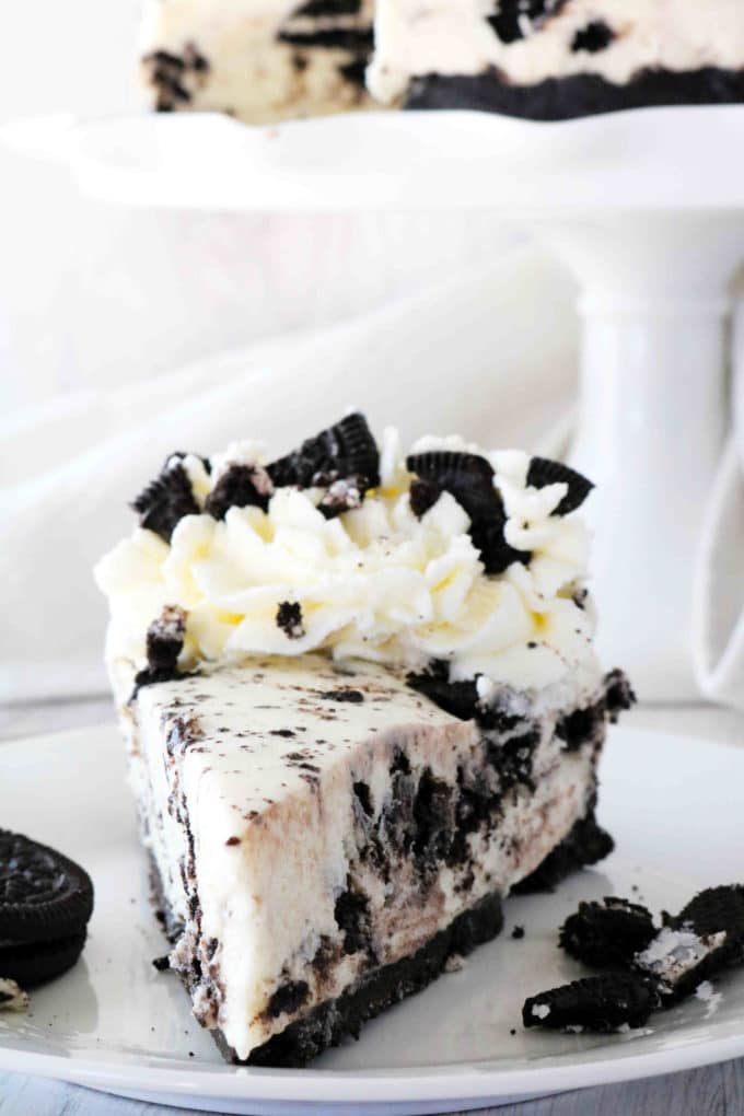 A slice of No-Bake Oreo Cheese on a plate with a cake stand behind it.