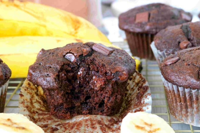 A muffin sitting on a cooling rack surrounding by banana slices and other muffins. It has a bite taken out of the center and looks very moist.