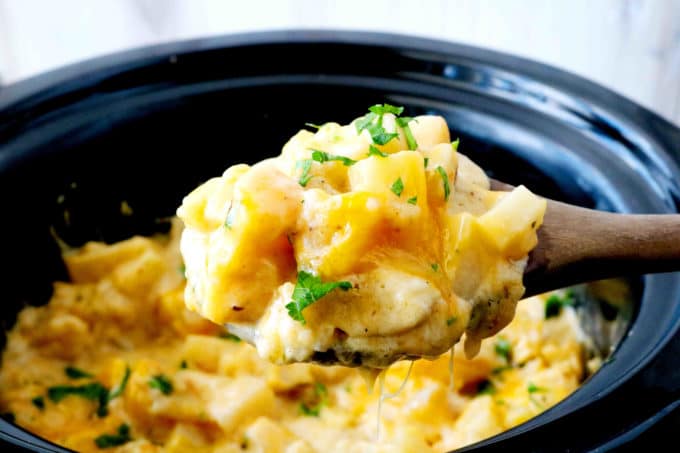 A wooden spoon full of cheesy crockpot potatoes being held over a crock-pot.