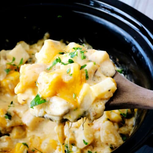A wooden spoon full of cheesy crockpot potatoes being held over a slow cooker.