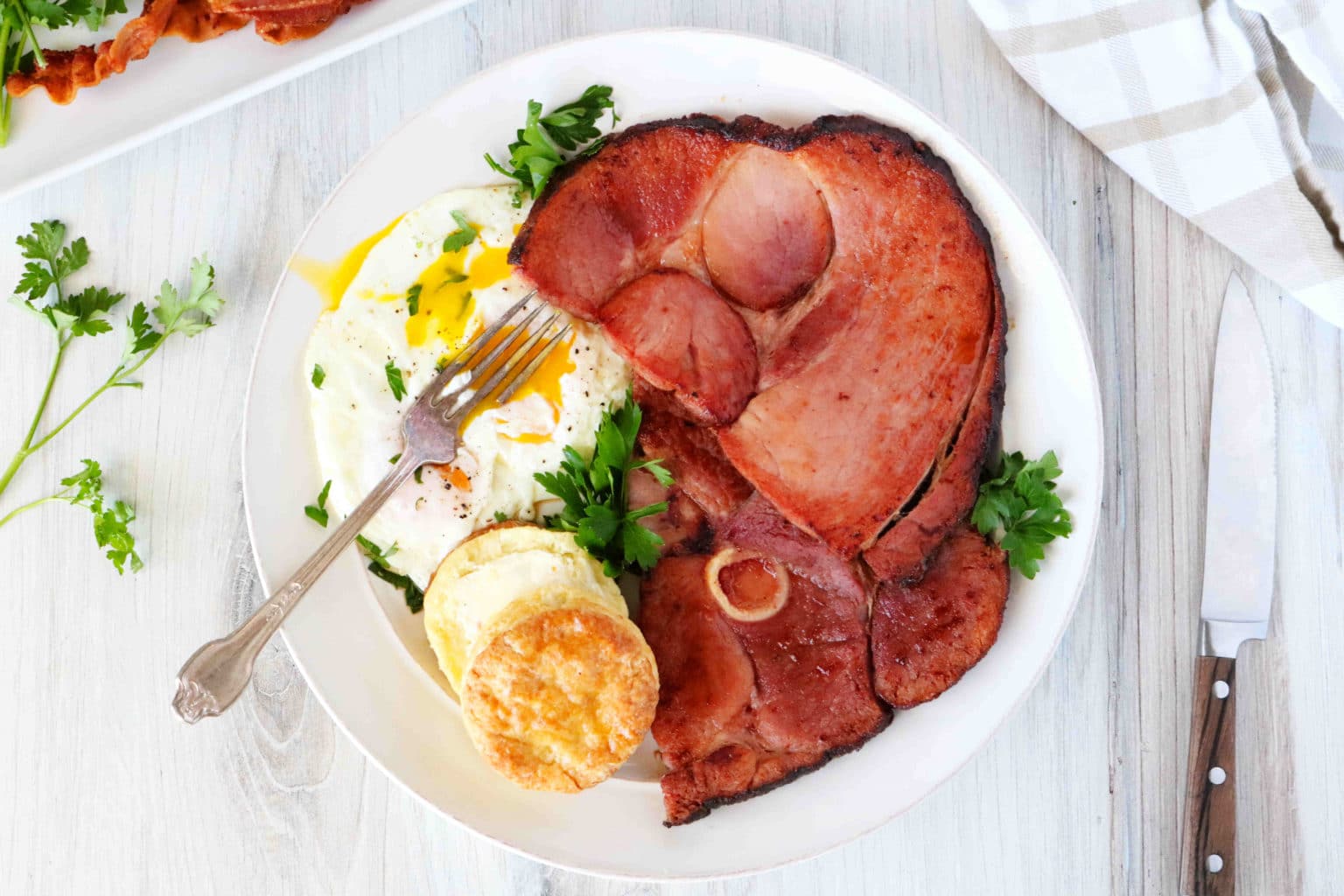 A bone-in ham steak on a plate with biscuits and fried eggs. 