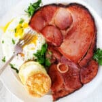 An overhead shot of ham steaks on a plate with an over-easy egg and a buttered biscuit.