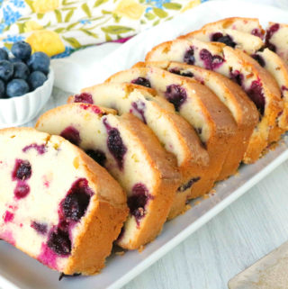 A close up shot of sliced blueberry pound cake on a platter with lemon and blueberries in the background.