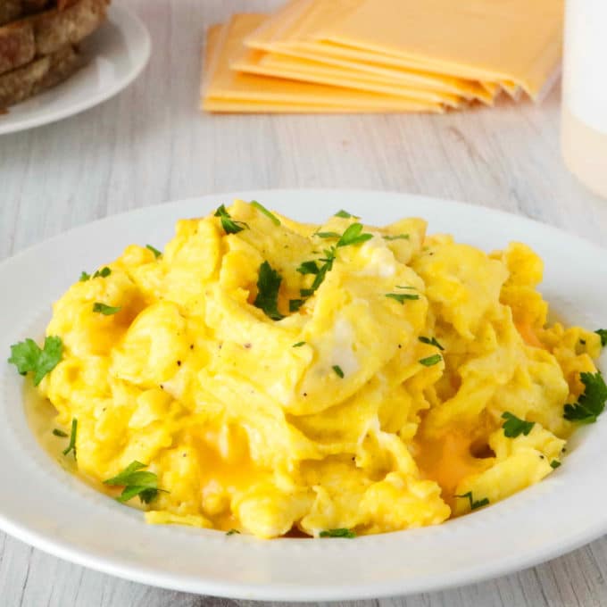 A plate of cheesy scrambled eggs with sliced cheese and toast behind it.