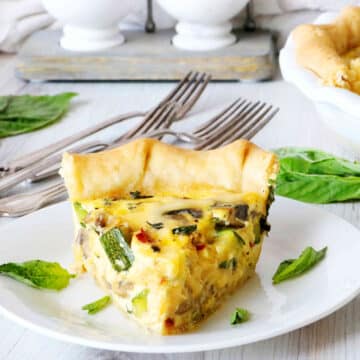 A slice of quiche sitting on a plate surrounded by basil leaves.