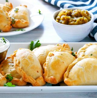 A platter of empanadas with a bowl of green chiles behind it.