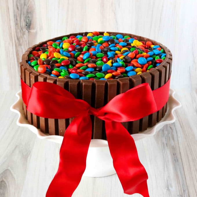 A front view of a chocolate cake, surrounded by KitKats, topped with M&Ms and tied with a red ribbon.