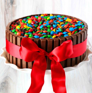 A front view of a chocolate cake, surrounded by KitKats, topped with M&Ms and tied with a red ribbon.