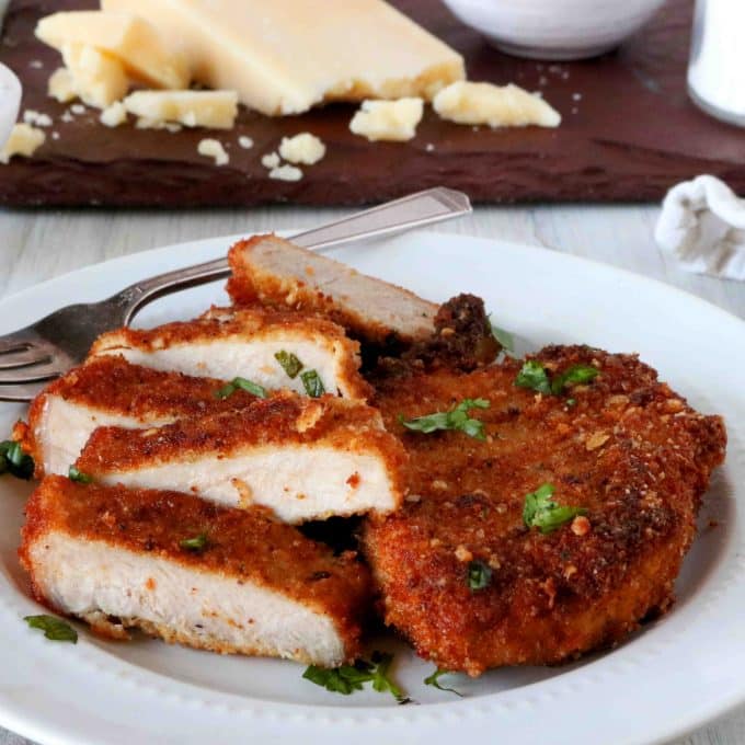 Parmesan Crusted Pork Chops with parsley sprinked over the top on a plate, cut into slices. There is a tray with parmesan cheese and herbs off to the side.