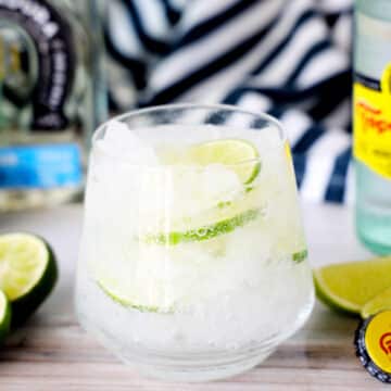 A Ranch Water Cocktail in a short glass with a bottle of tequila, lime wedges, and topo chico behind it.