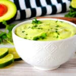 A bowl of avocado crema with a cilantro garnish and avocados, limes, and a striped napkin in the background.