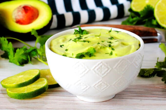 A bowl of avocado crema with a cilantro garnish and avocados, limes, and a striped napkin in the background.