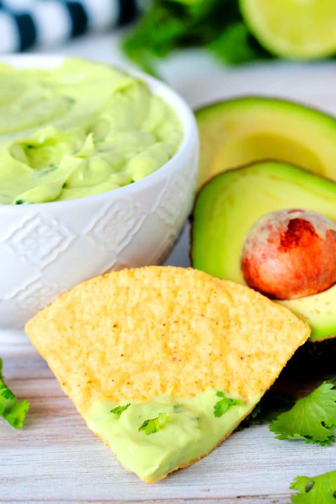 A chip dipped in Avocado Crema with avocados and a bowl of dip in the background.