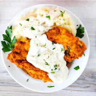 Chicken fried chicken on a plate topped with gravy and mashed potatoes on the side.