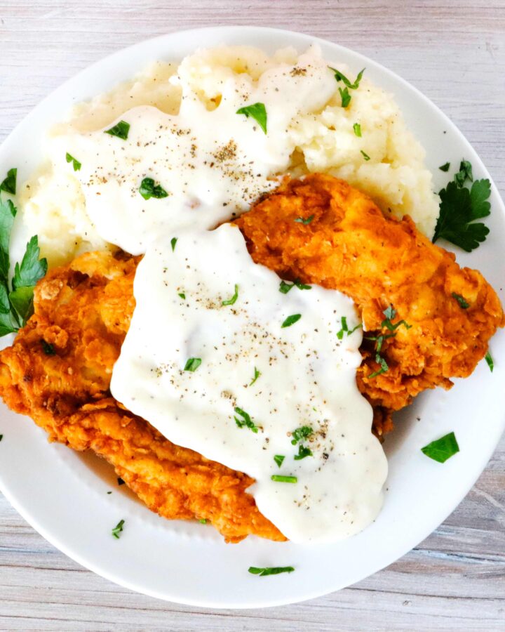 Chicken fried chicken on a plate topped with gravy and mashed potatoes on the side.