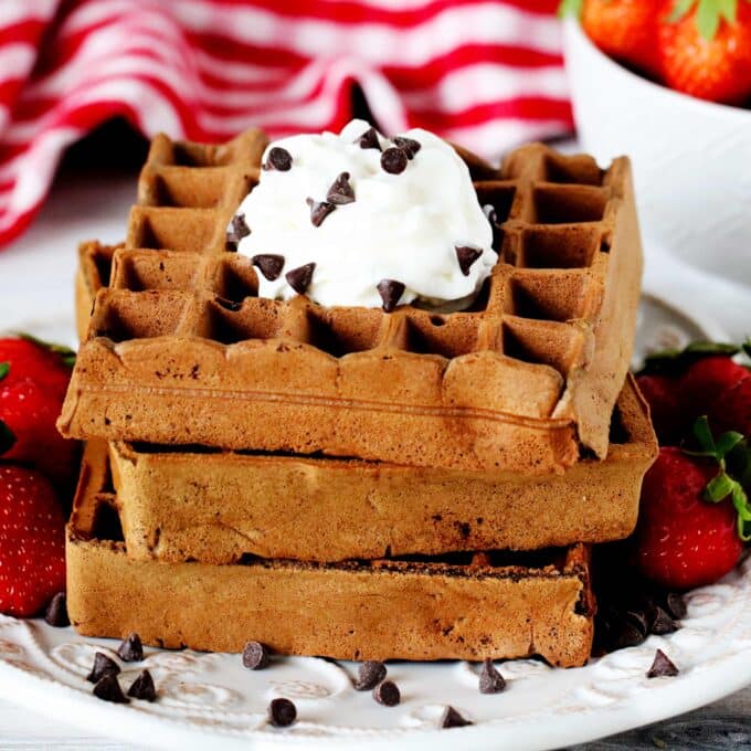 A stack of chocolate waffles on a plate surrounded by strawberries and chocolate chips with a checkered towel and a bowl of strawberries behind it.