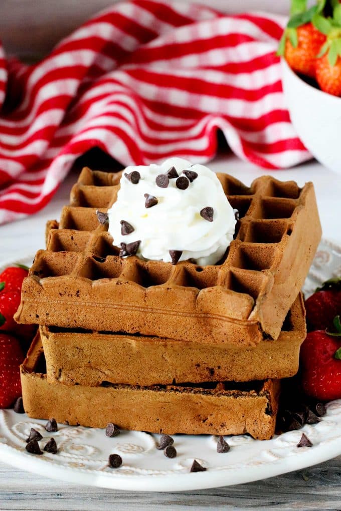A stack of chocolate waffles on a plate surrounded by strawberries and chocolate chips with a checkered towel and a bowl of strawberries behind it.