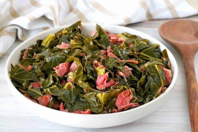 A white bowl full of collard greens with shredded ham hock with a spoon off to the side and towel in the background.