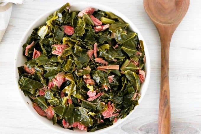 An overhead shot of a white bowl full of collard greens with shredded ham hock and a spoon off to the side and towel in the background.