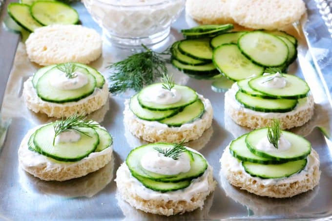 A tray of assembled cucumber sandwiches with a dollop of dill sauce on top and a sprig of dill for garnish
