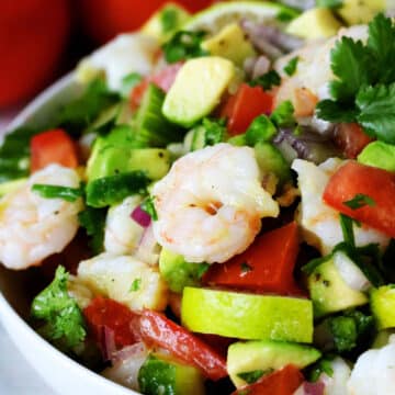 A close up shot of shrimp salad in a white bowl with tomato, avocado, lime, and cilantro.
