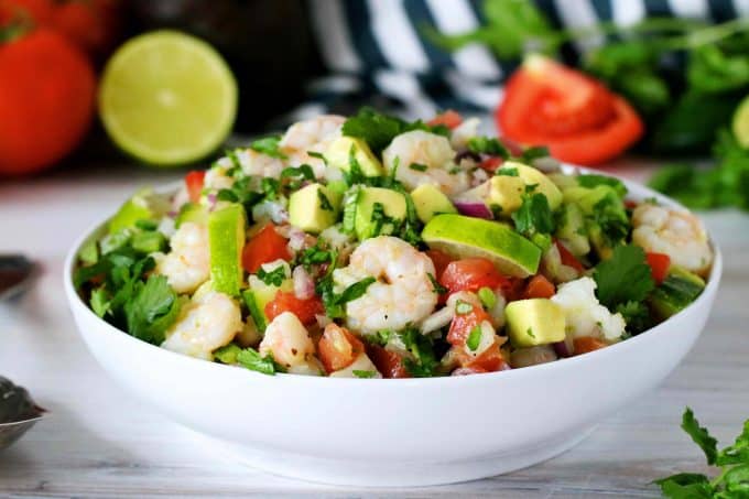 Shrimp Avocado Salad in a white bowl with tomatoes, limes, and avocado in the background.