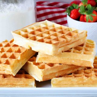 A square image of a stack of waffles on a platter with waffle mix and strawberries behind it.
