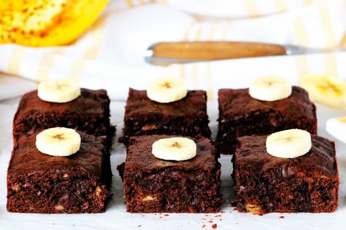 Banana brownies cut into squares with a slice of banana on top with a knife and a banana in the background.