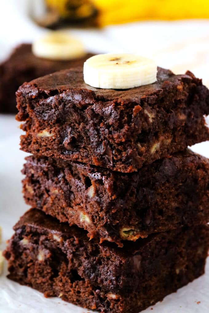 Three chocolate banana brownies stacked on top of one another with a sliced banana on top.