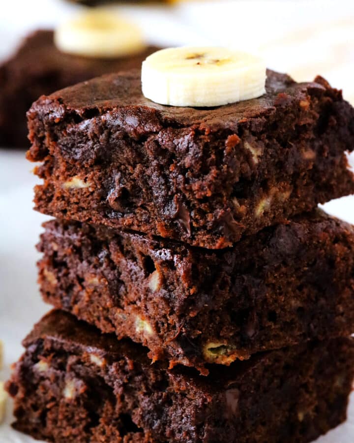 Three chocolate banana brownies stacked on top of one another with a sliced banana on top.