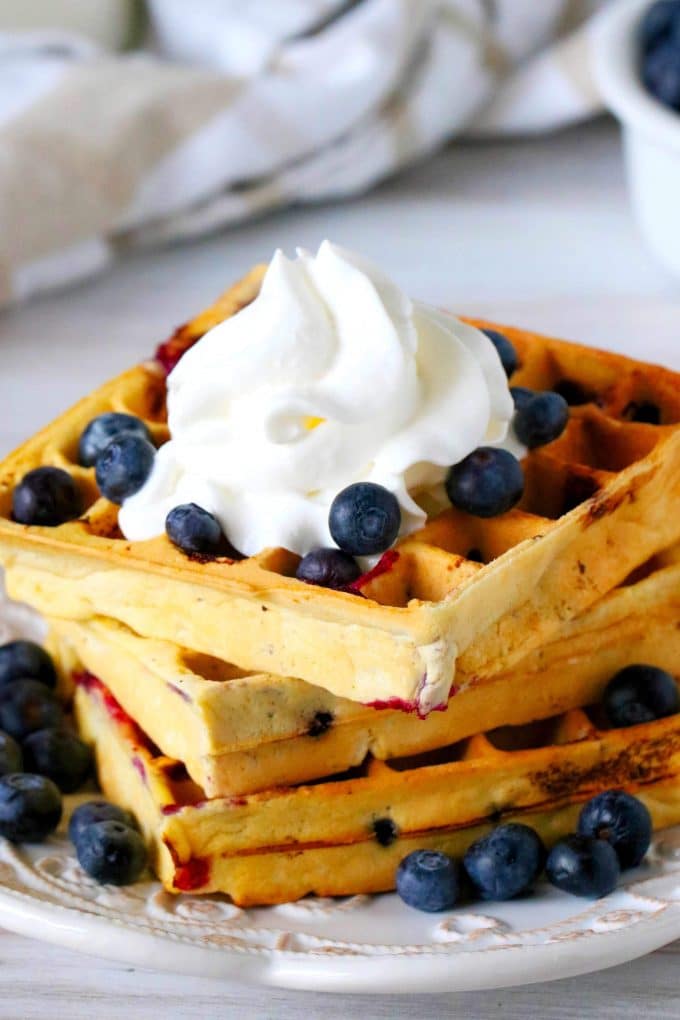 Blueberry Waffles on a plate with blueberries surrounding it and whipped cream on the top.