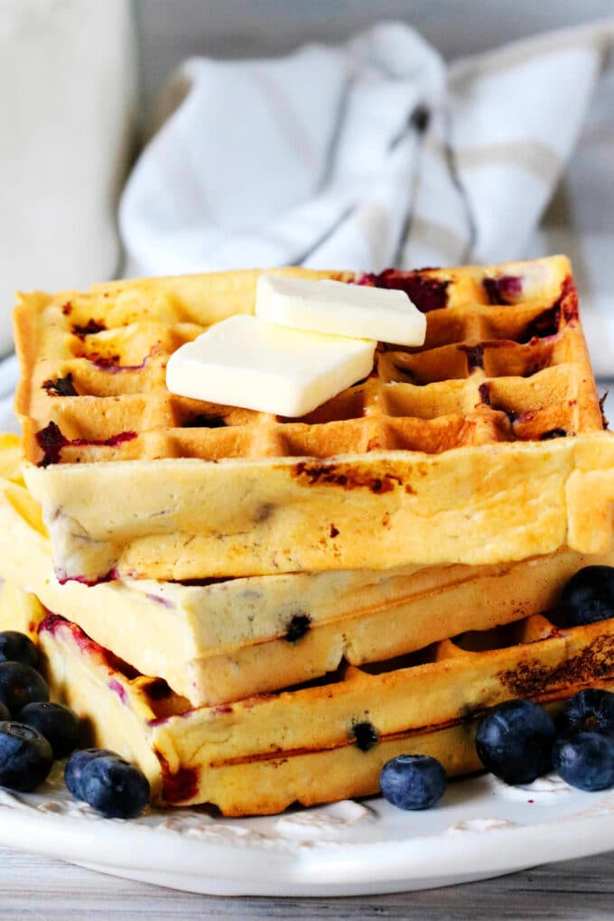A stack of Blueberry waffles on a plate with blueberries surrounding it and butter on top.