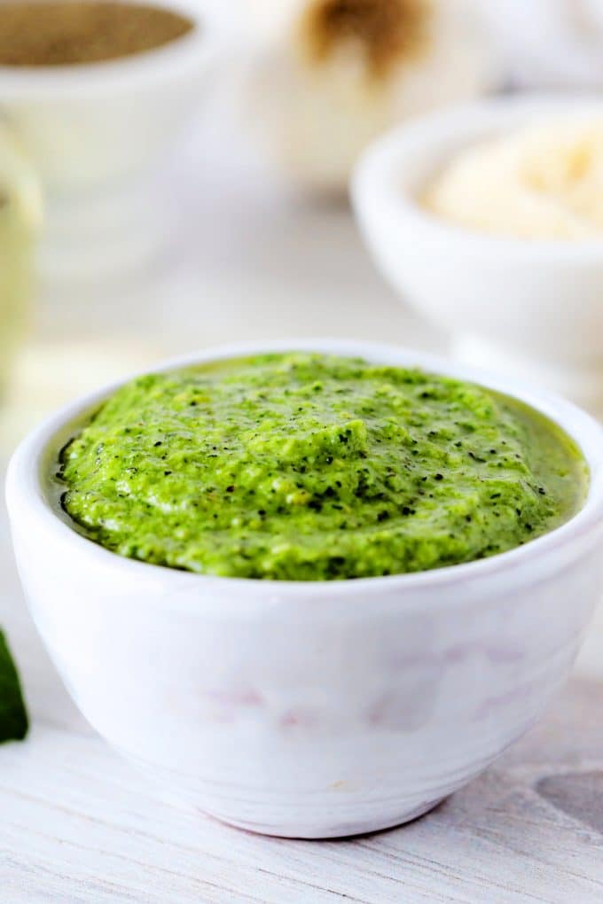 A bowl of creamy pesto with parmesan, oil pepper, and garlic in the background.