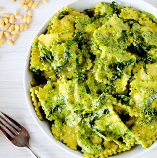 An overhead shot of ravioli tossed in a creamy pesto sauce with a fork and a scatter of pine nuts off to the side.