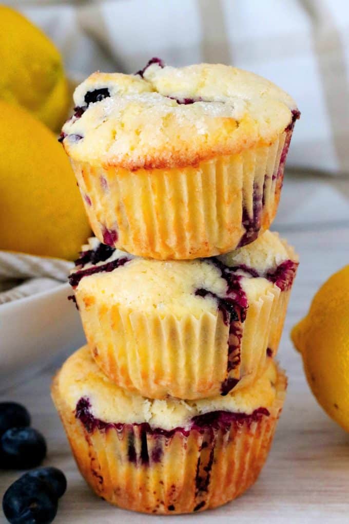 Three lemon blueberry muffins stacked on top of one another with lemons and blueberries off to the side. 