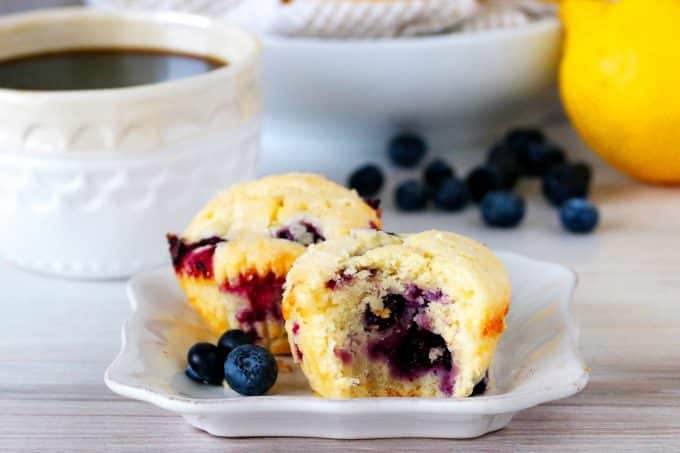 Lemon blueberry muffins on a plate with a cup of coffee behind them.