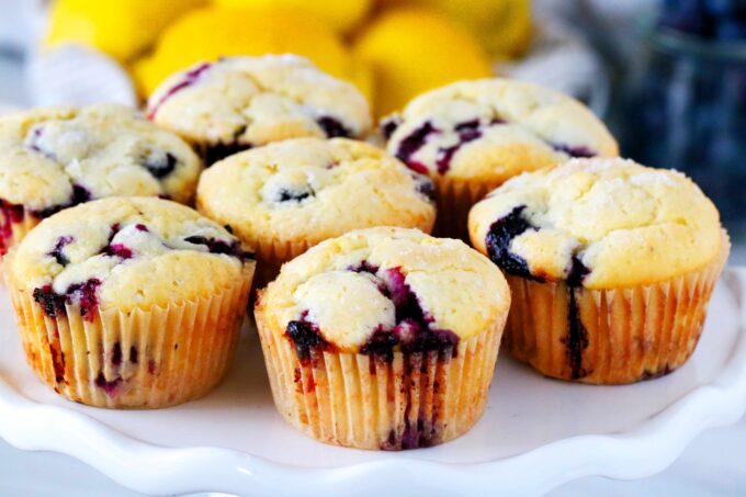 A cake stand with lemon blueberry muffins on top of it and lemons in the background.