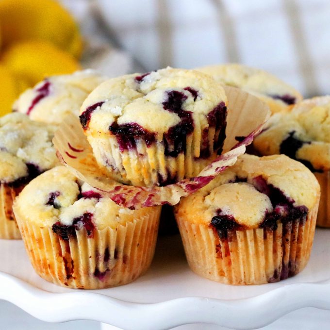 A cake stand with lemon blueberry muffins on top of it and lemons in the background.