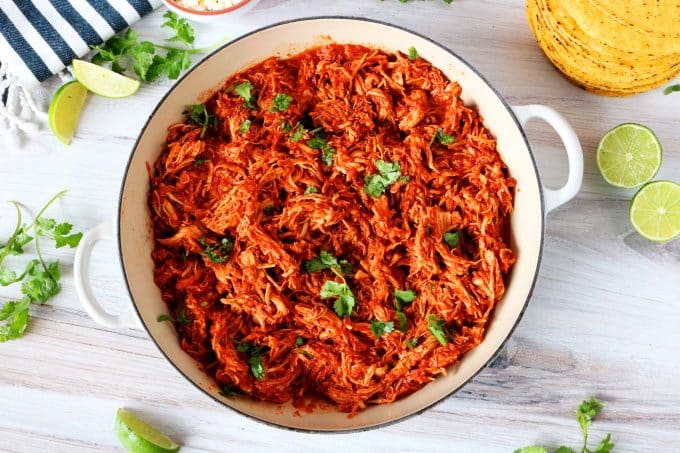 An overhead shot of chicken tinga in a braising dish with limes, tostadas, and cilantro surrounding it.