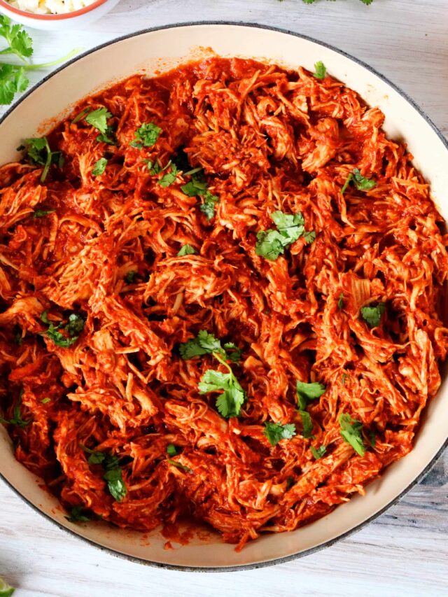An overhead shot of chicken tinga in a braising dish with limes, tostadas, and cilantro surrounding it.